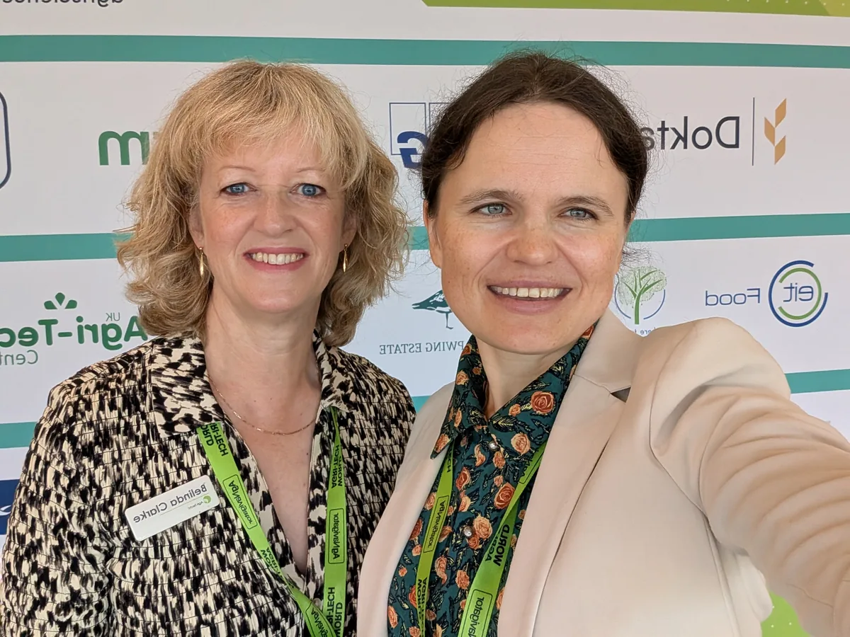 Dr. Maryna Kuzmenko in a selfie with Belinda Clarke, in a patterned jacket and green lanyard, in front of a sponsor wall.