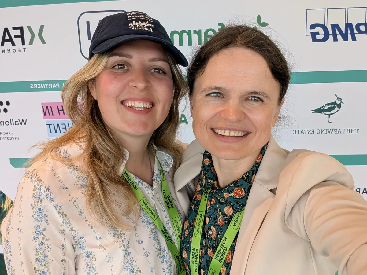 Dr. Maryna Kuzmenko in a selfie with Emma Chapelhow, in a cap and a floral blouse, in front of a sponsor wall.
