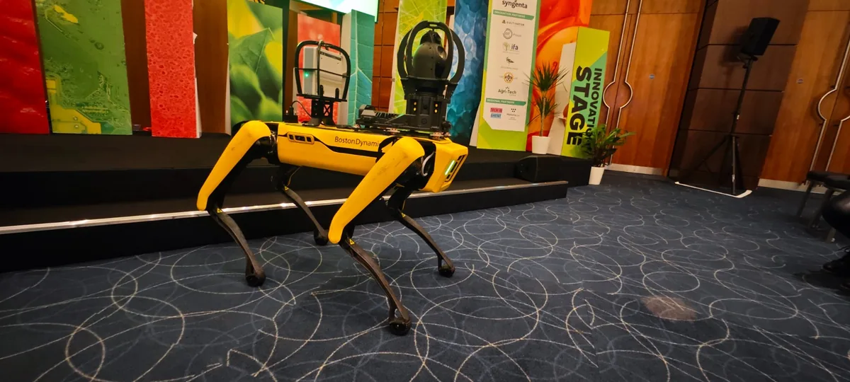 A yellow Spot quadruped robot standing on the patterned carpet of the summit hall, in front of the stage backdrop.