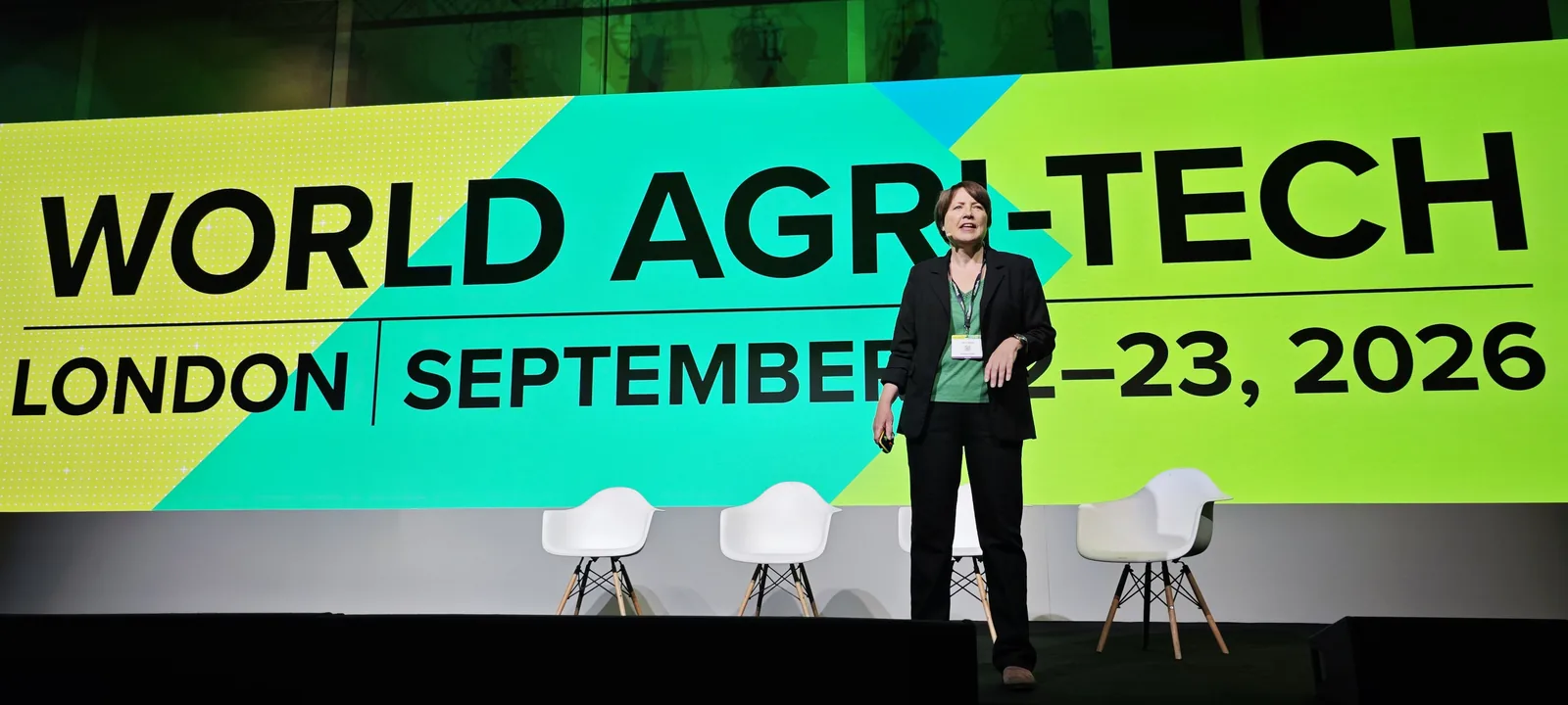 Jane Byrne standing alone on a wide stage in front of a bright green and yellow backdrop reading World Agri-Tech, London, September 22 to 23, 2026, with two white chairs beside her.