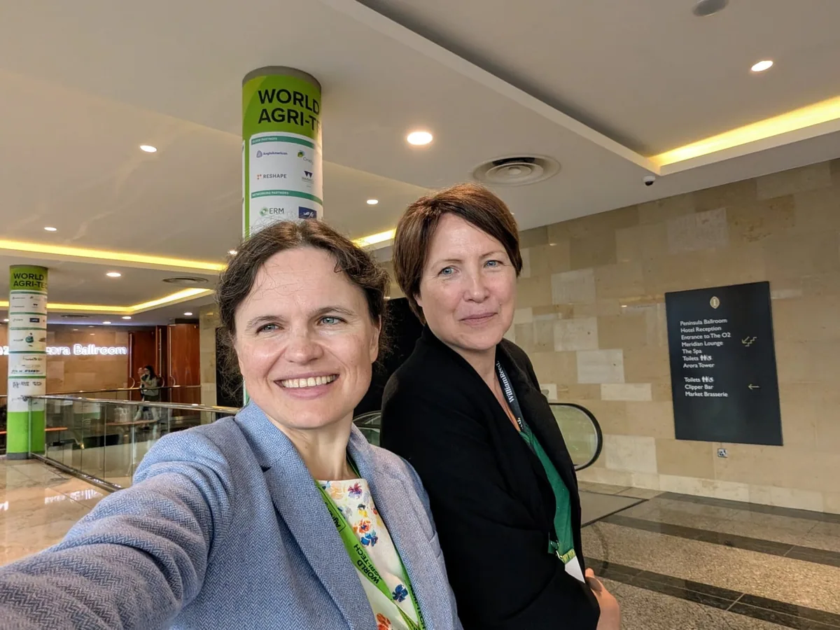Dr. Maryna Kuzmenko and Jane Byrne, both wearing green lanyards, in a selfie beside a World Agri-Tech banner in a hotel lobby.