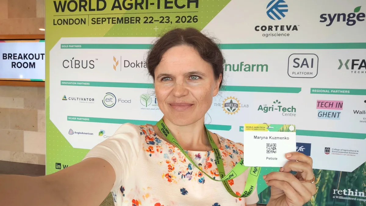 Dr. Maryna Kuzmenko holding up her delegate badge, in a selfie in front of the World Agri-Tech London, September 22 to 23, 2026 partner wall.