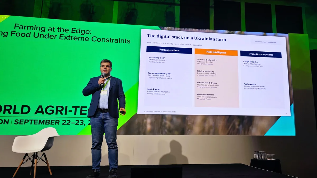 Volodymyr Pugachov speaking on the summit stage with a microphone, beside a slide titled Farming at the Edge: Growing Food Under Extreme Constraints, showing the digital stack on a Ukrainian farm.