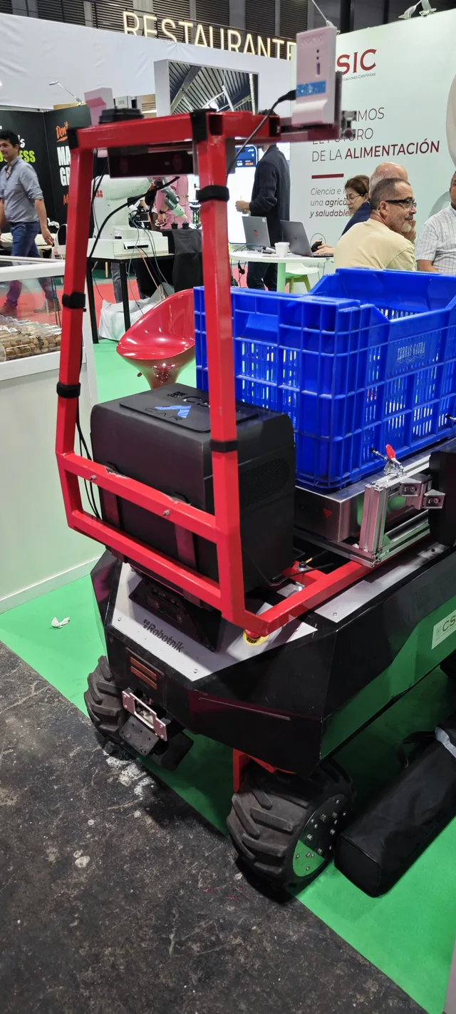 A compact wheeled robot with a red frame carrying a blue produce crate, on a stand.