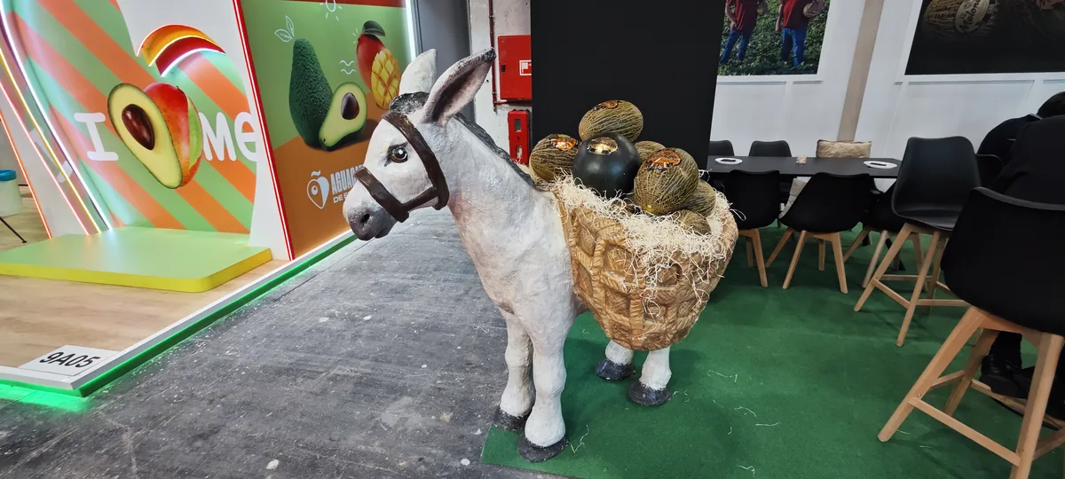 A life-size model donkey carrying baskets of produce, on green carpet in front of an avocado-themed stand.