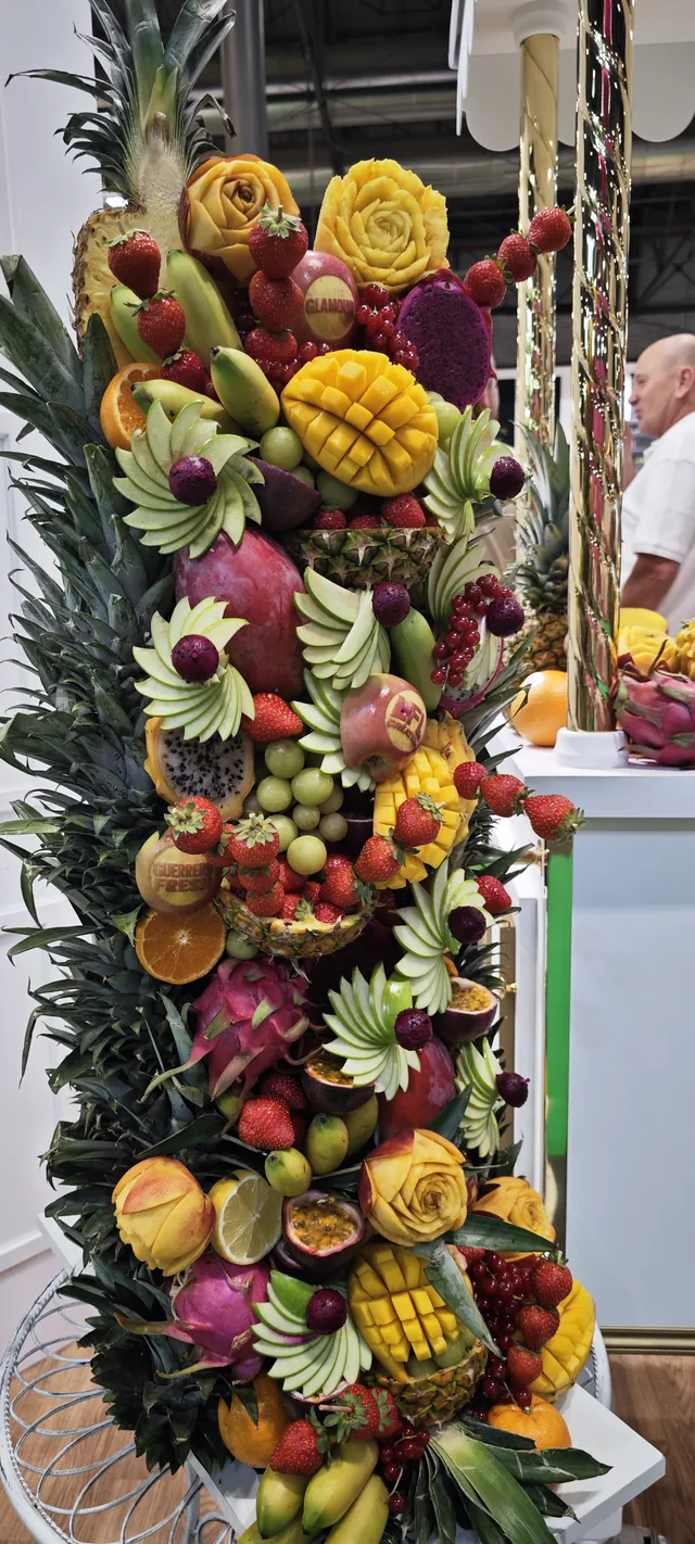 A tall pillar built entirely of fresh fruit: pineapples, dragon fruit, melons, grapes and leaves, stacked from floor to ceiling.