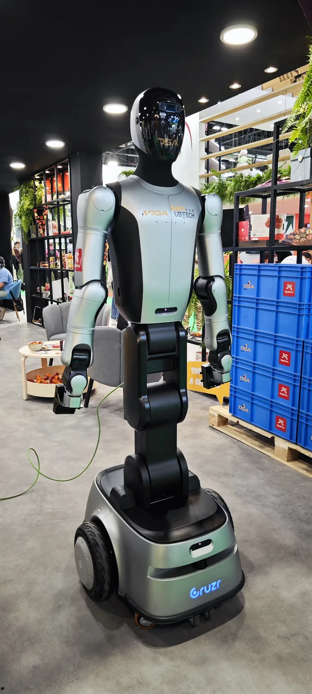 A white humanoid robot on a wheeled base in an exhibition aisle, beside crates of produce.