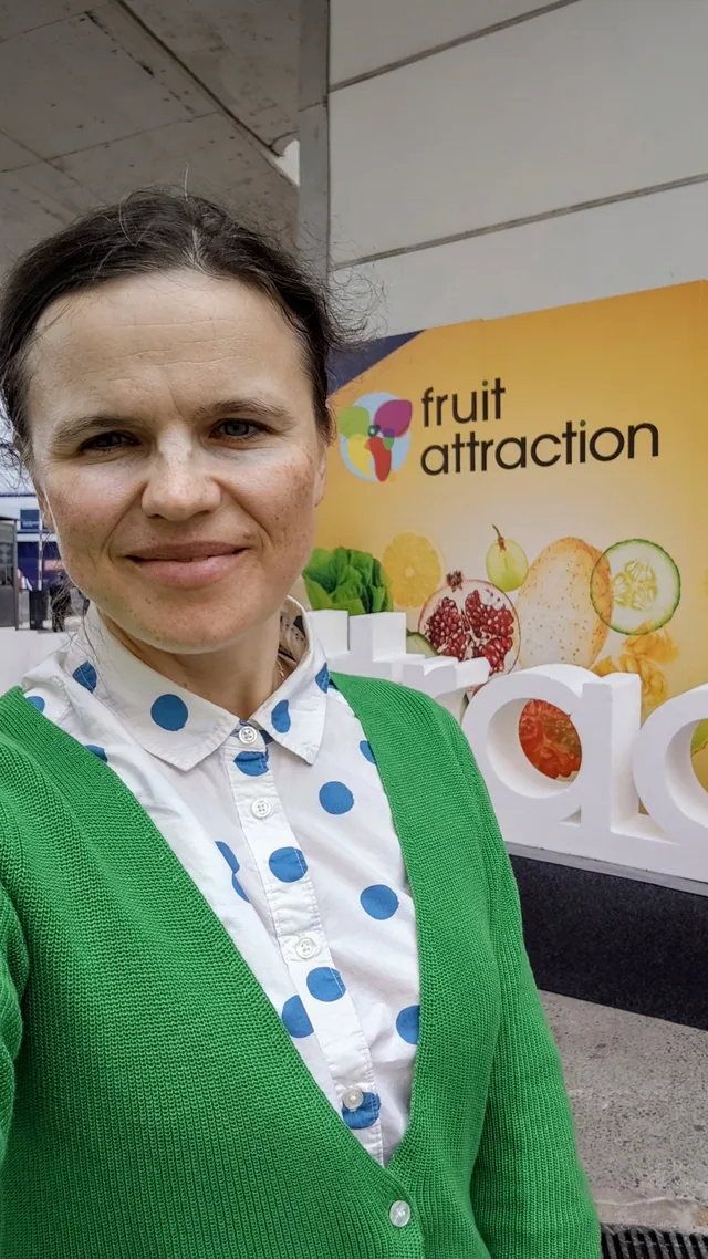 Dr. Maryna Kuzmenko in a green cardigan and polka-dot blouse, in a selfie in front of the Fruit Attraction entrance banner.