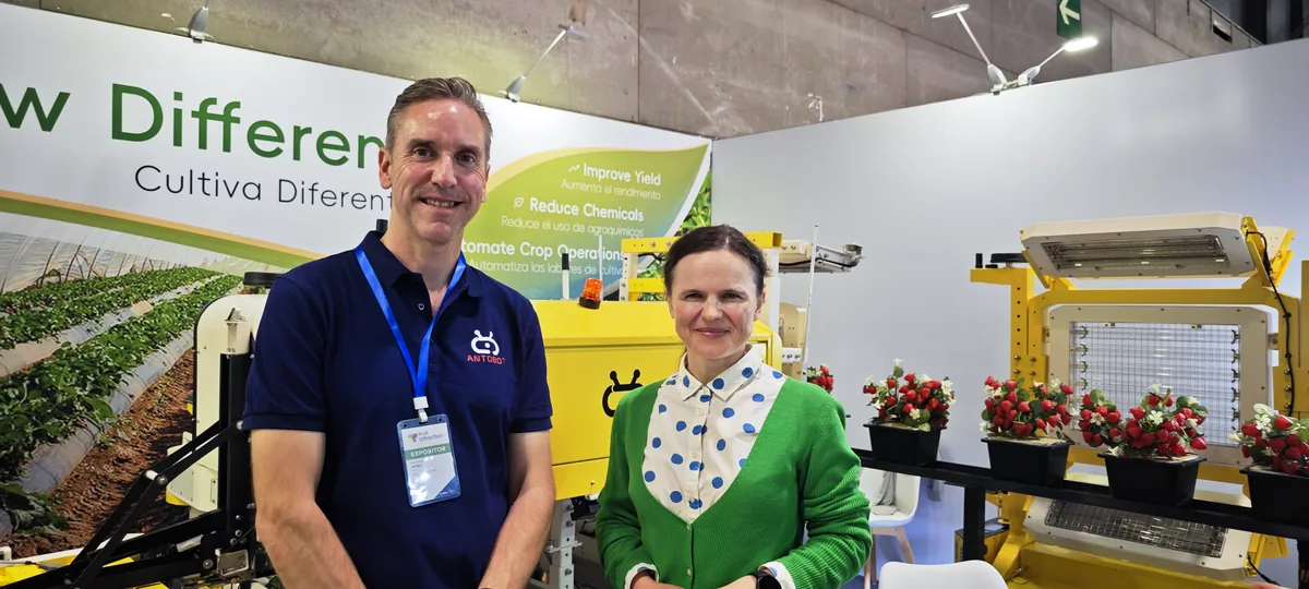 Dr. Maryna Kuzmenko and Marc Jones standing in front of an Antobot stand with yellow field robots and a wall of strawberry plants.