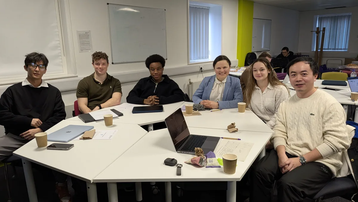 Six people seated around joined seminar tables with laptops, notebooks and coffee cups, in a university seminar room with a whiteboard behind them.
