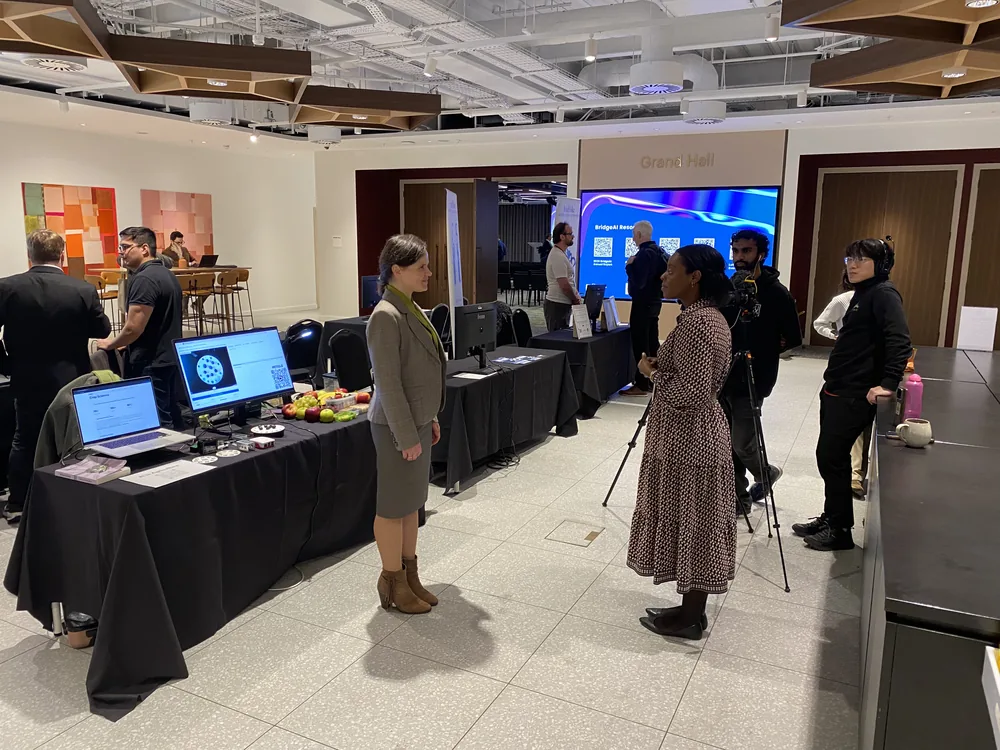Dr. Maryna Kuzmenko standing beside a demonstration table of apples and screens showing apple quality analysis, in a hall with a BridgeAI slide behind her.