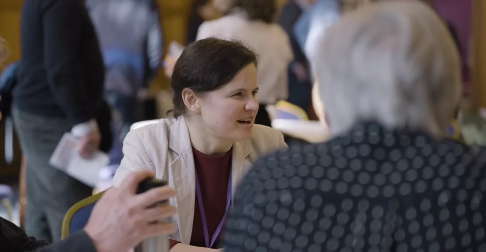 Dr. Maryna Kuzmenko talking with another delegate across a table in a busy conference hall.