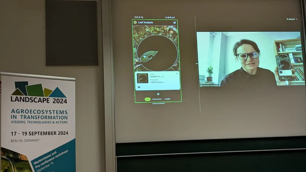 A lecture-hall screen showing Dr. Maryna Kuzmenko on a video call holding up a phone, beside a projected leaf-area measurement, next to a banner reading Landscape 2024, Agroecosystems in Transformation, 17 to 19 September 2024, Berlin, Germany.