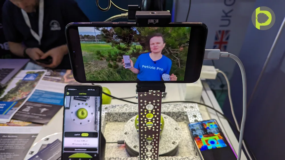 An exhibition stand rig: a phone on a mount above apples on a Petiole measuring board, a second phone reading an area of 1.94 square centimetres, and a third showing a thermal image.