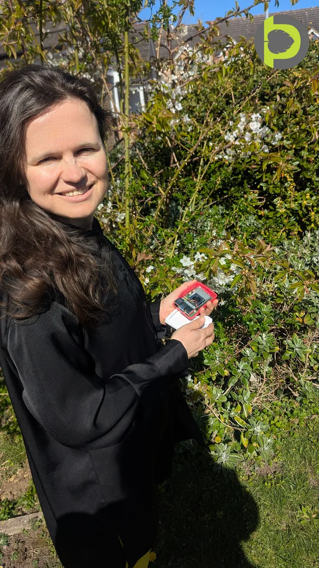 Dr. Maryna Kuzmenko holding a Raspberry Pi and camera module in a red case, in a garden beside a flowering hedge.