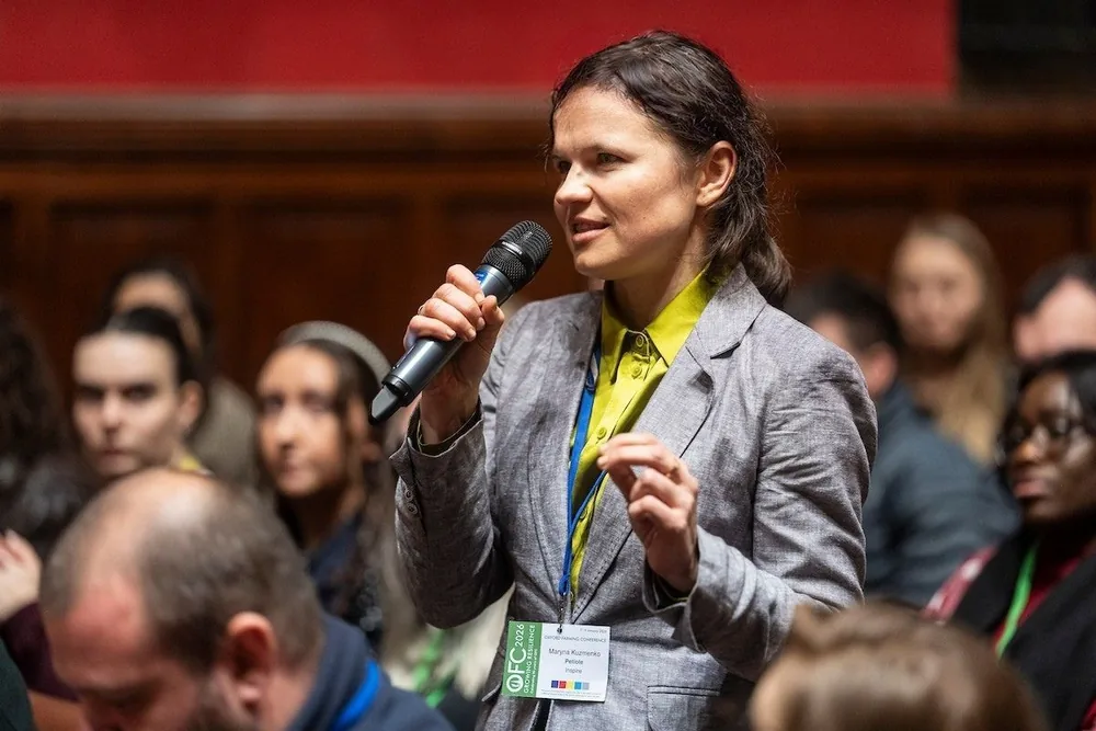 Dr. Maryna Kuzmenko speaking into a handheld microphone, standing among a seated audience in a wood-panelled hall.