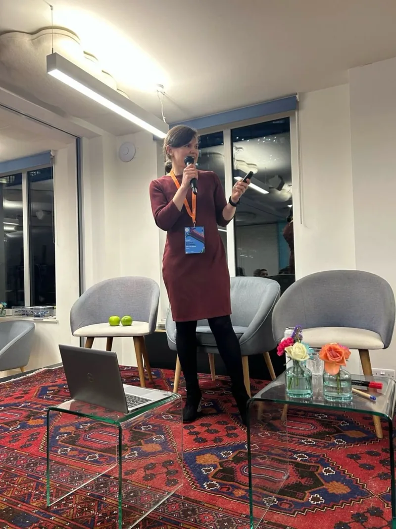 Dr. Maryna Kuzmenko standing and speaking into a microphone in a meeting room, a laptop open on a low table beside her.