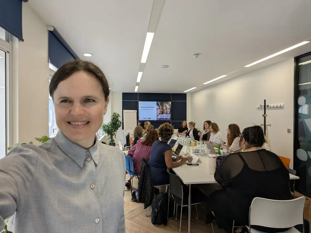 Dr. Maryna Kuzmenko in the foreground of a bright meeting room, a group working around tables behind her beneath a slide reading Women in Innovation Community Forum.